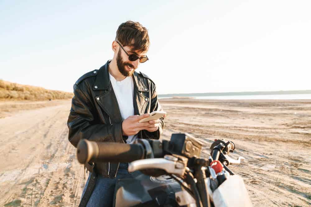 smiling young man sitting on motorcycle comparing insurance quotes on phone