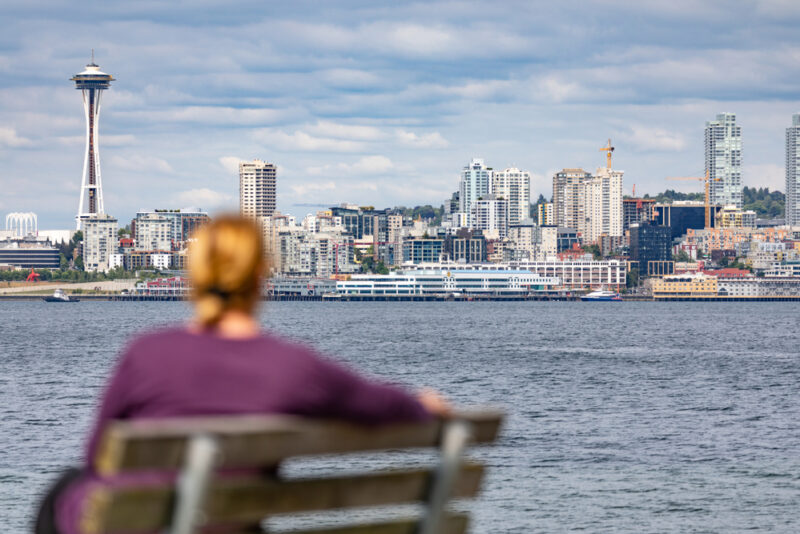 Woman sits on bench gazing across water at Seattle Space Needle - cheap car insurance in Washington.