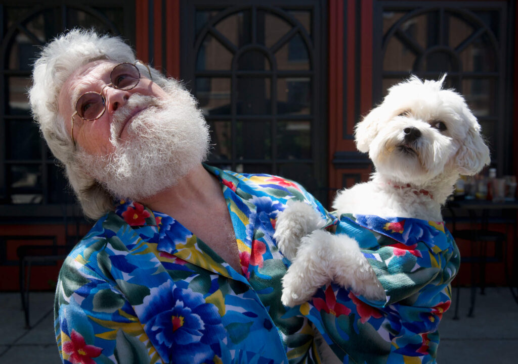 Older man with white hair and his small dog with white hair looking in opposite directions with matching tropical shirts.