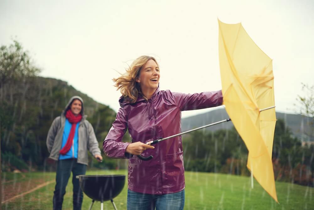 Man and woman outside grilling when rainstorm kicks up, woman trying to open umbrella - cheap umbrella insurance in Washington.