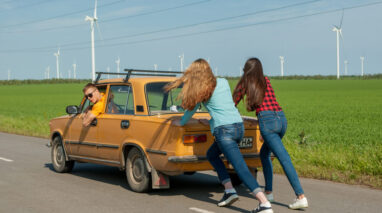 two girls pushing little yellow car that is broken down with a guy steering.