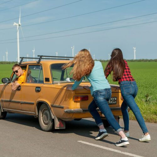 two girls pushing little yellow car that is broken down with a guy steering.