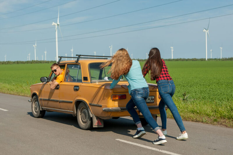 two girls pushing little yellow car that is broken down with a guy steering.